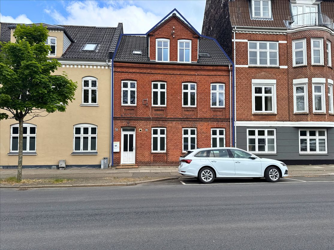 En hvid bil parkeret foran en rød bygning med flere vinduer og én dør, og en træstamme udenfor