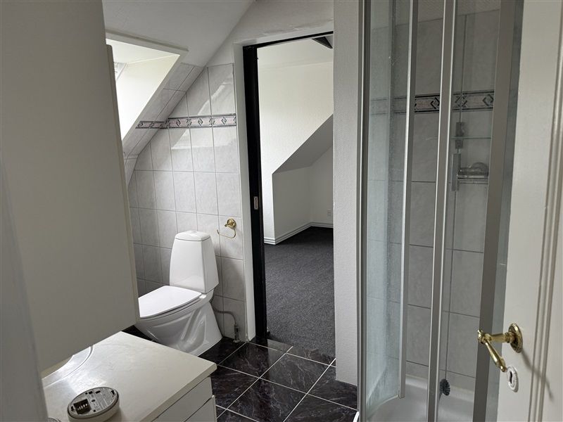 Badrum med hvidt toilet og brusekabine med glasdør placeret i et loftsrum med skrå loft