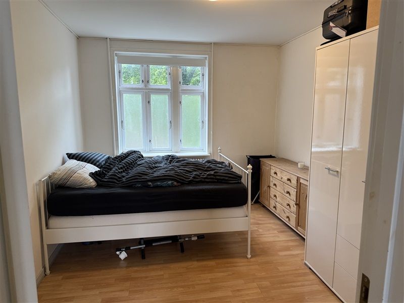 Et soveværelse med hvidt sengetøj og en hvid metalramme. Høj garderobe med flere skuffer og en svart kuffert på toppen.