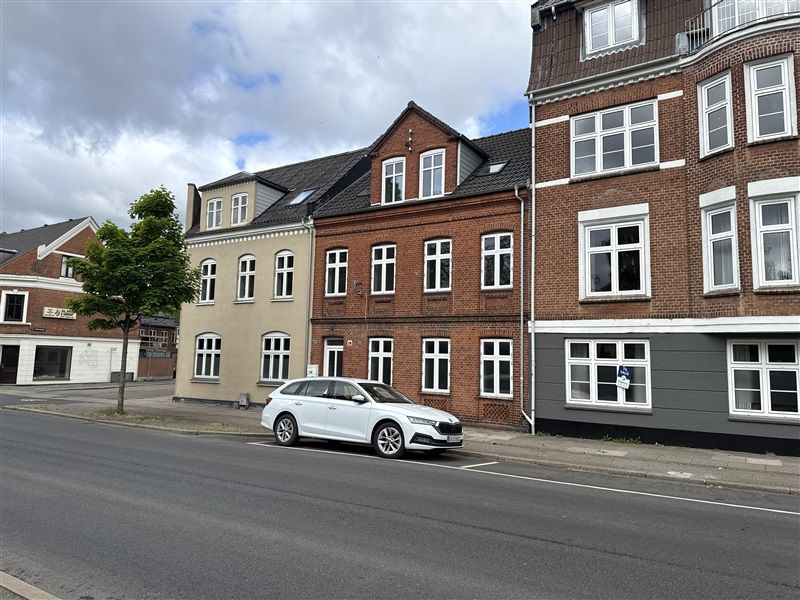 Bilparkeret hvid stationcar på gaden foran tre etagers røde og grå ejendomme med flere vinduer og en gul væg.