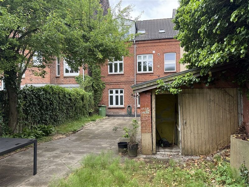 Ejendom med rækkehus og garage, udstyret med skur og have med planter, sammen med træer og grønne affaldscontainere