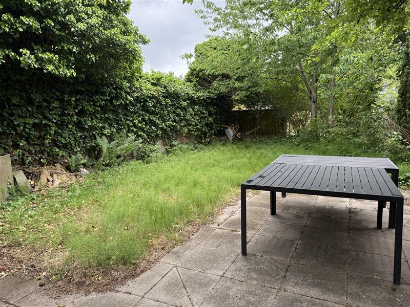 En sort bord er anbragt på en terrasse i en grøn have med træer og græs. En trækurv ligger bagved på græsset.