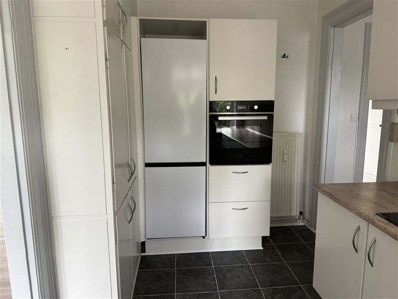 Komplet indrettet køkken med hvide vægge, køleskab og indbygget ovn samt hvide skabe med håndtag og lysegrå bordplade