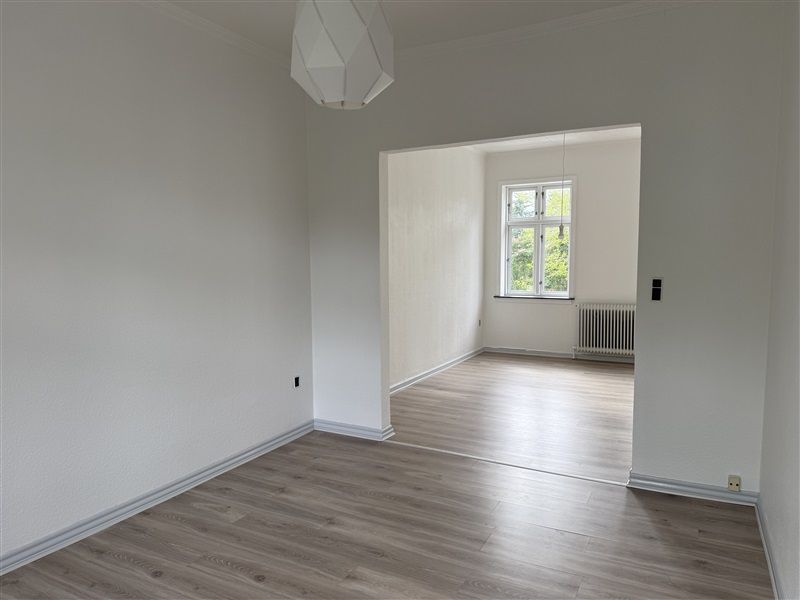 En tom lejlighed med hvidt vægmaleri og lysegul vinylgulv og en åbning til et soveværelse med vindue og radiator