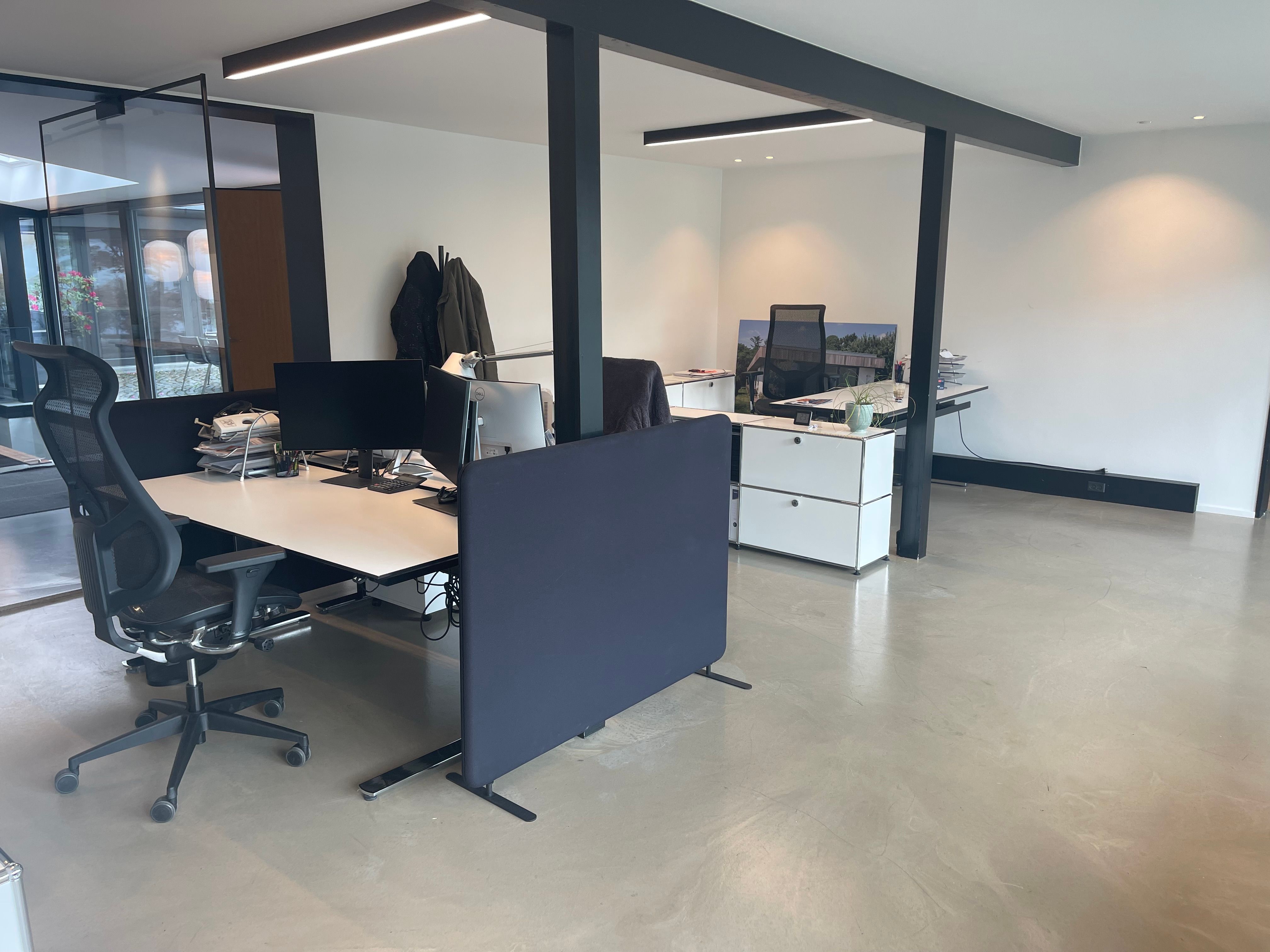 Et moderne kontor med lyse vægge og to skriveborde med monitorer og plante i hjørnet