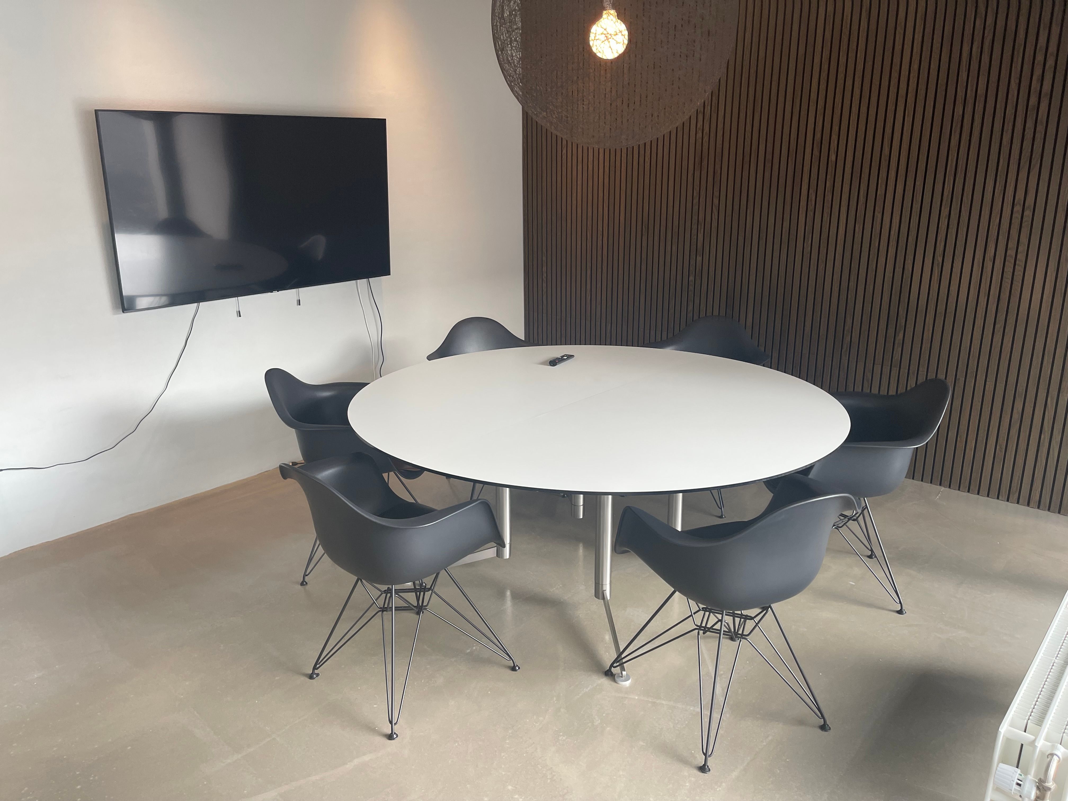 Hvid rund konferencebord med seks stole og en stor tv-skærm på væggen i en co-working space