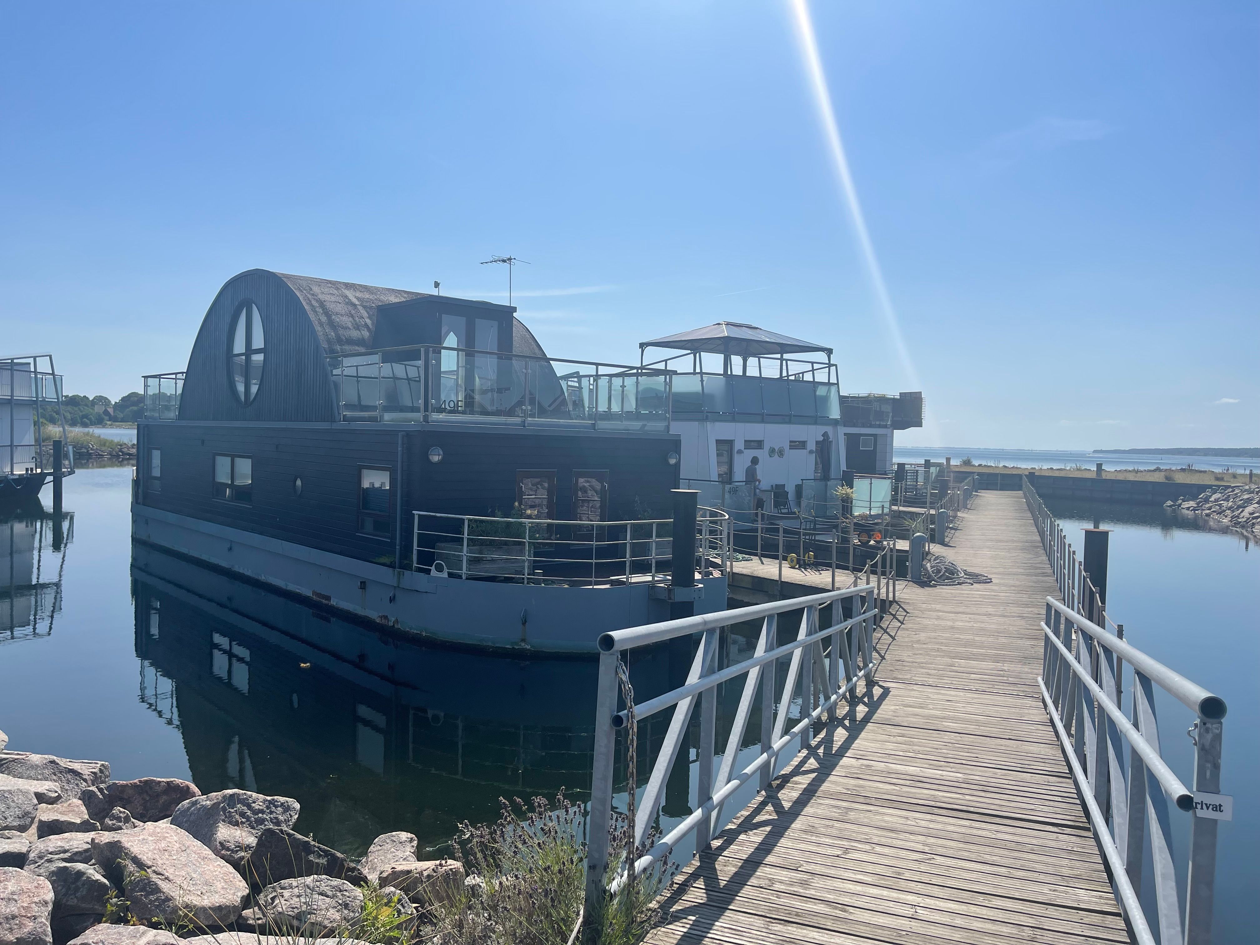 En nyrenoveret husbåd med to dæk, der er forankret i en havn med en gangbro og udsigt over vandet til landskabet i øst.