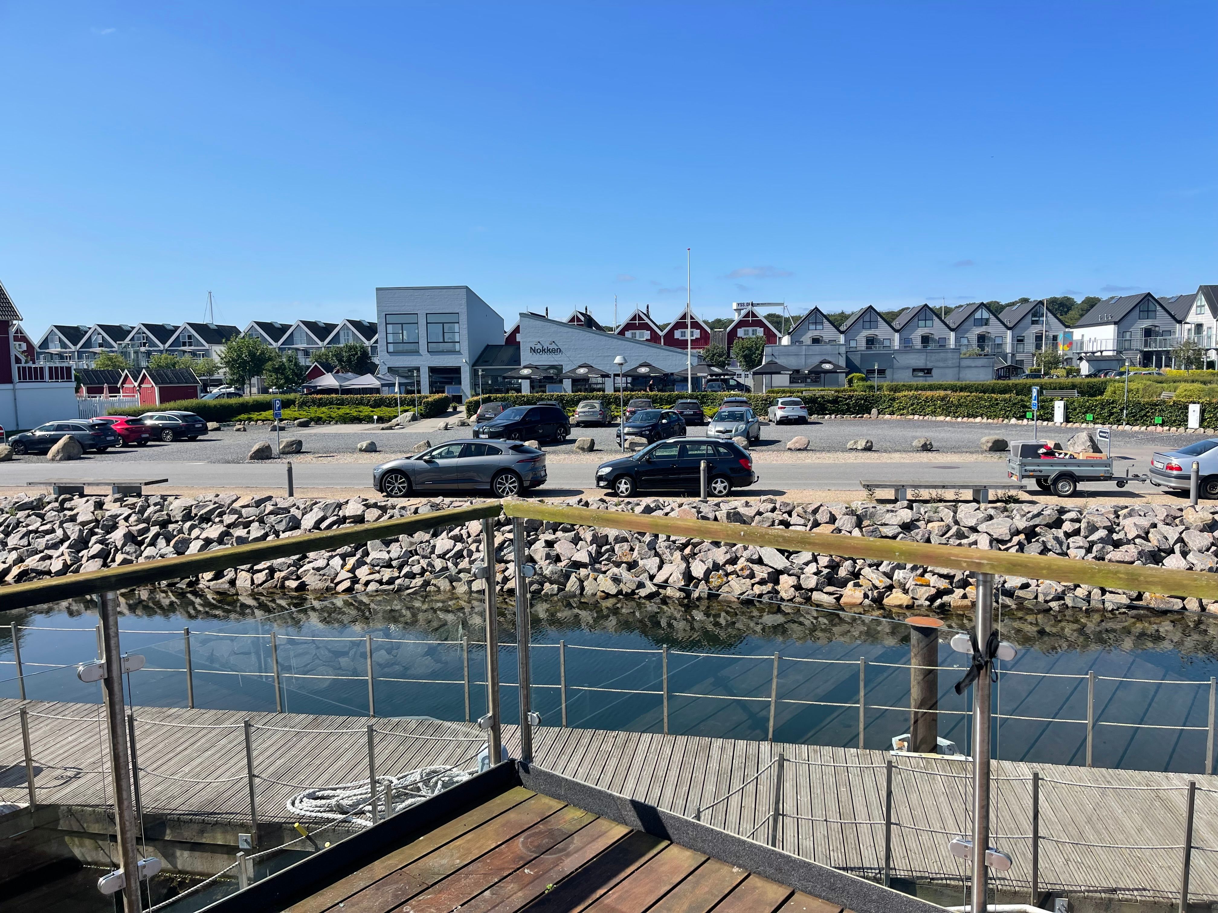 Mange biler parkeret i parkeringsplads nær stranden, en udsigt over vandet og træbro, en række huse med forskellige farver