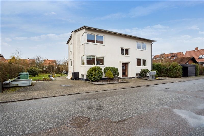 Et hvidt to-etagers hus med store ruder og træindgang, omgivet af grønne buske og en grønt overdækket affaldscontainer i