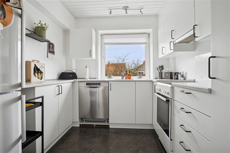 Køkken med hvide vægge, lysekroner, hvide skabe, komfur, ovn, opvaskemaskine, loft og vinduer med udsigt til grønne træer