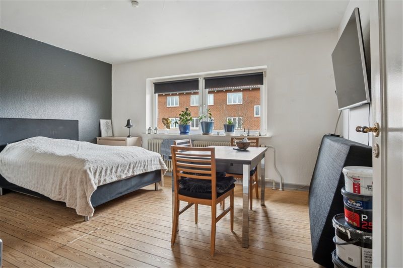 Et soveværelse med dobbeltseng og skrivebord med to stole, en tv-skærm monteret på væggen og vinduer med gardiner og