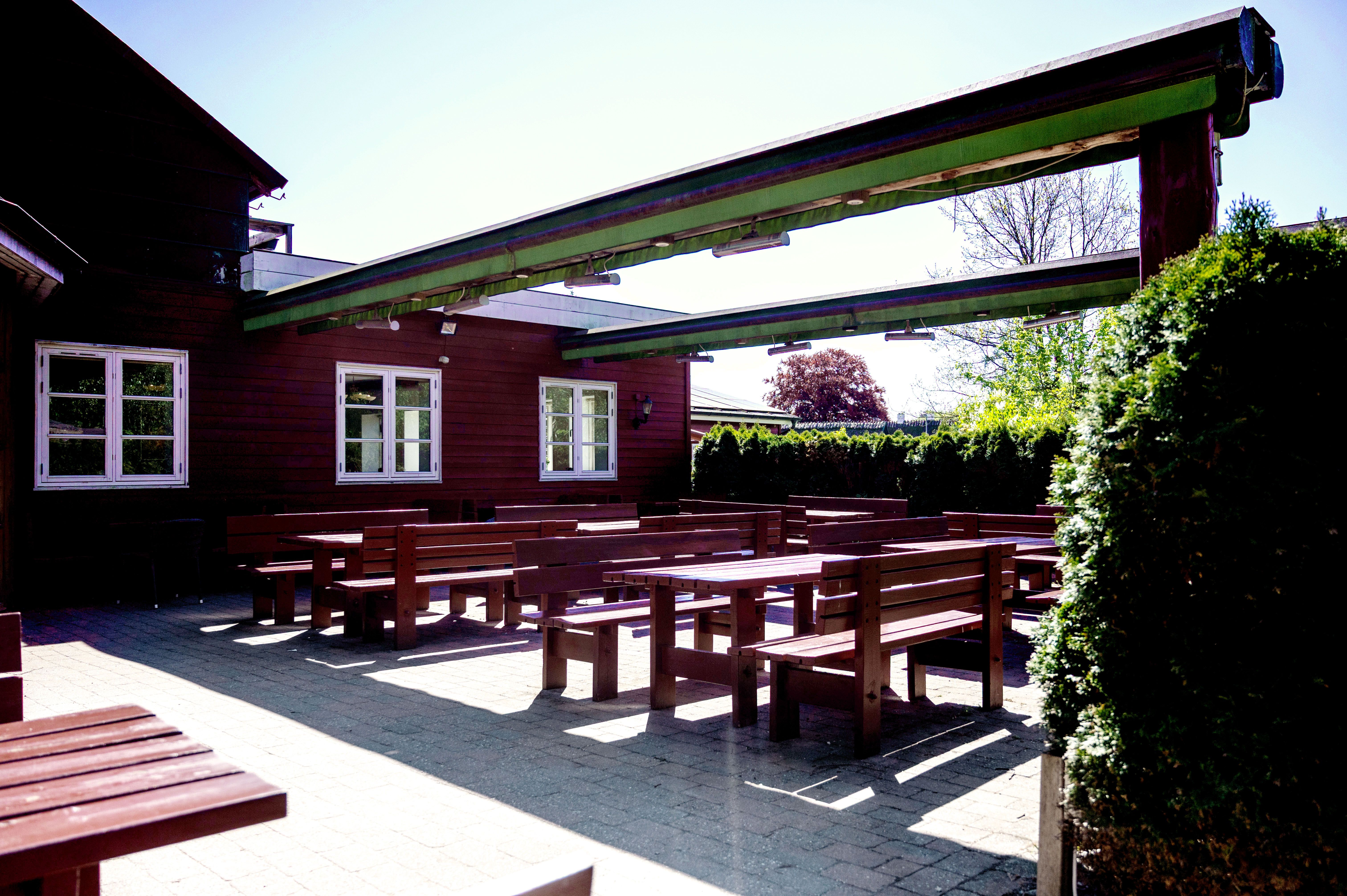 Træborde og bænke i en udendørs spisestue, et træpergola og buske i baggrunden