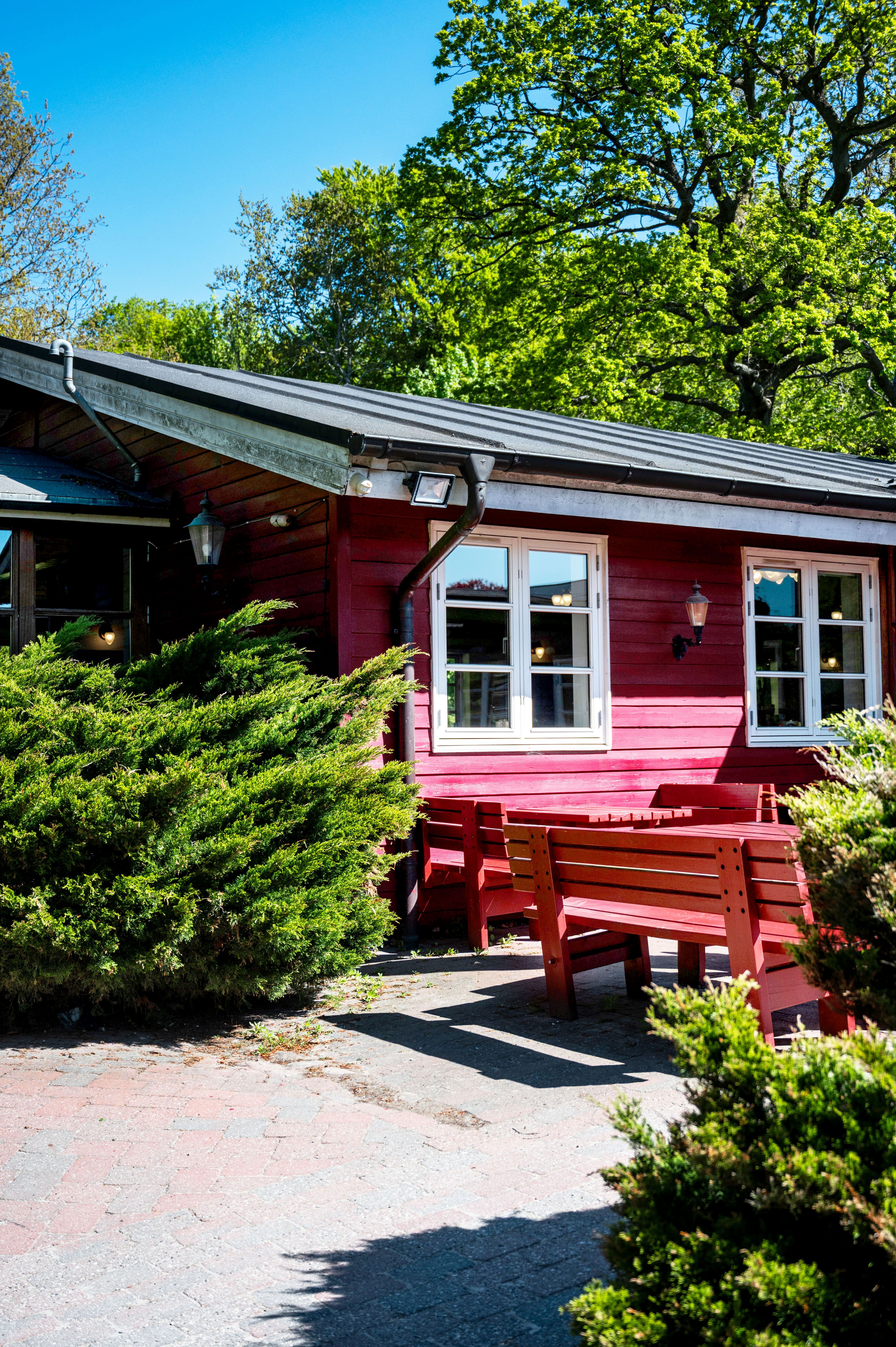 En rødmalet bygning med terrasse og udendørs møbler, alt foret med buske, under en sommerdag med mange træer