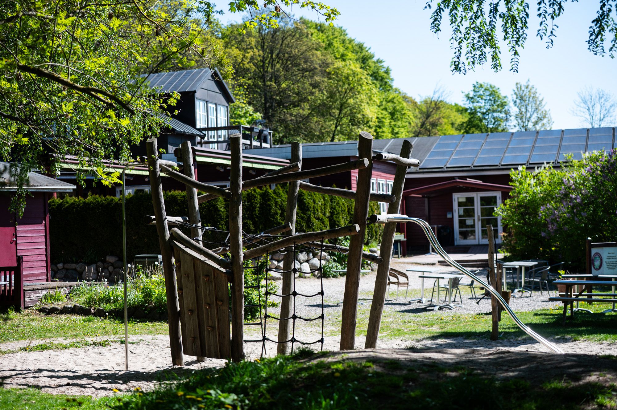 Solcellepaneler på tag og parkerede biler i baggrunden. Udendørs legeplads med klatrevægge og sandbane. Bord og stole i