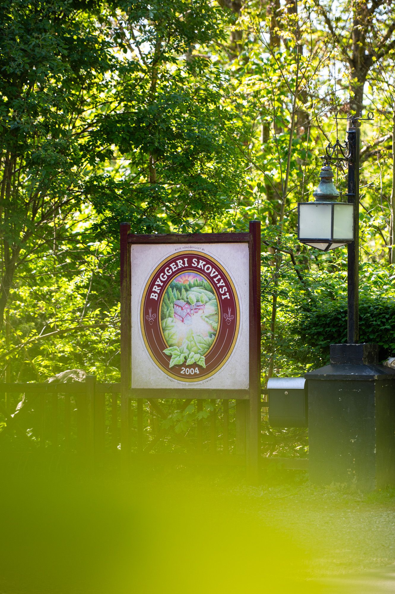 Et stort skilt med logo og tekst står ved siden af en lampe og skov i dagslys