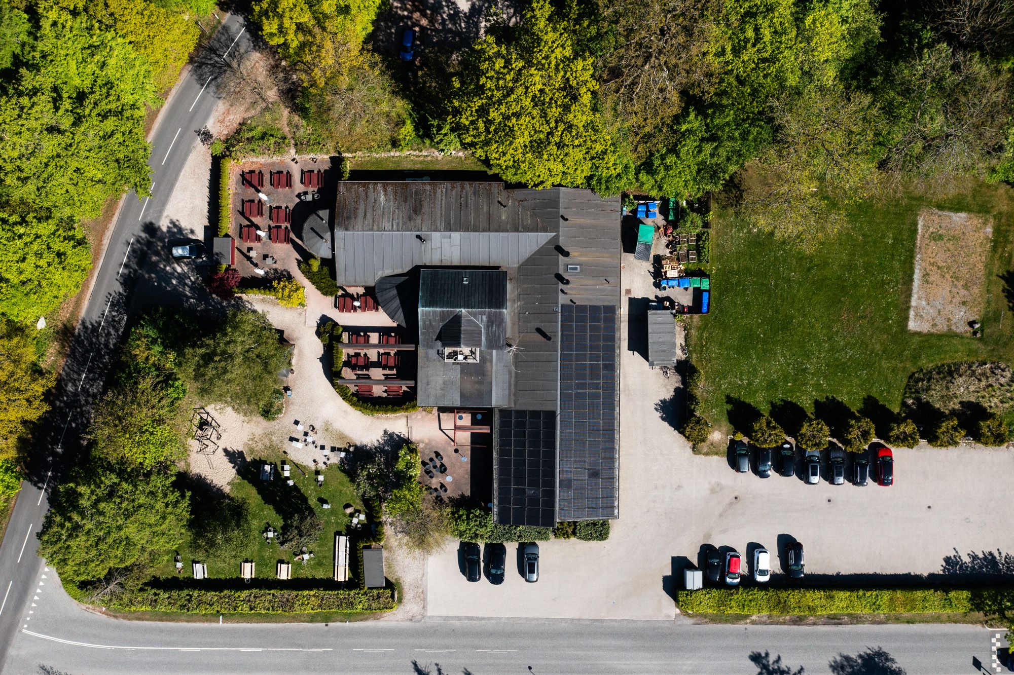 Et moderne, solcellepanelsdækket bygning med parkeringsplads og gæsteborde. Bygningen har to indgange og er omgivet af