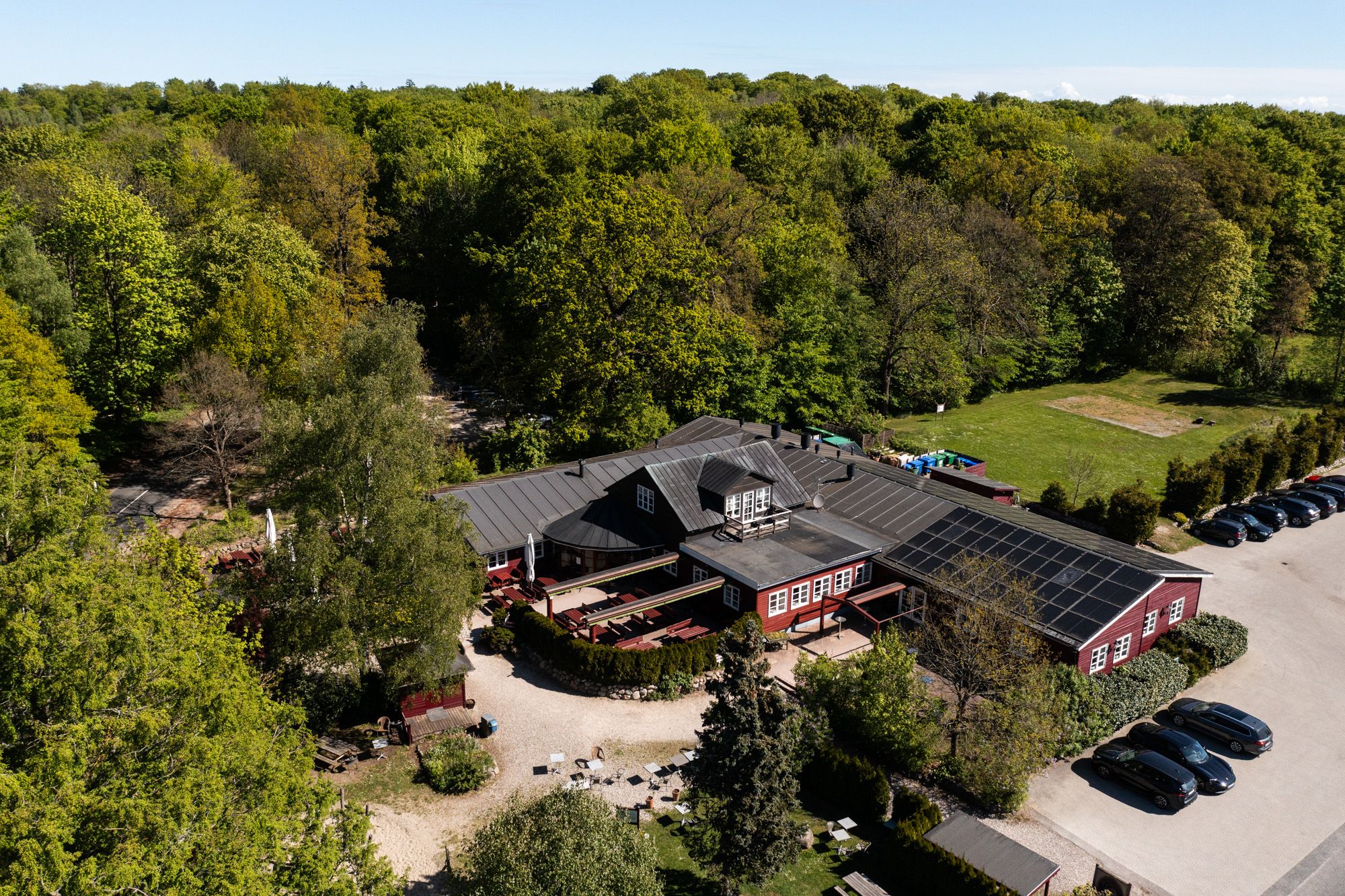 Bygning med solpaneler og parkeringsplads, parkeret biler og træer med grønne løv. Bænke og borde placeret udenfor.