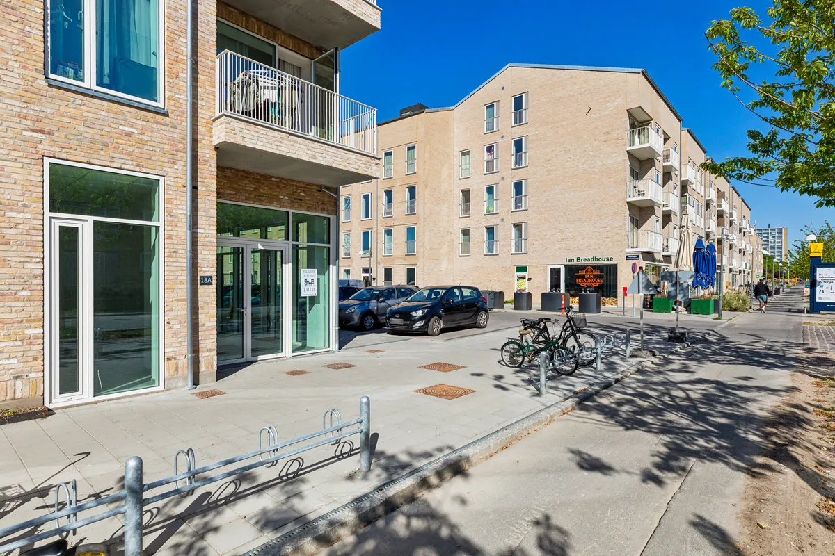 To højhusbygninger med balkoner og lejemål på Ruten 18A. Parkerede biler og cykler samt facade med skilt for Julian