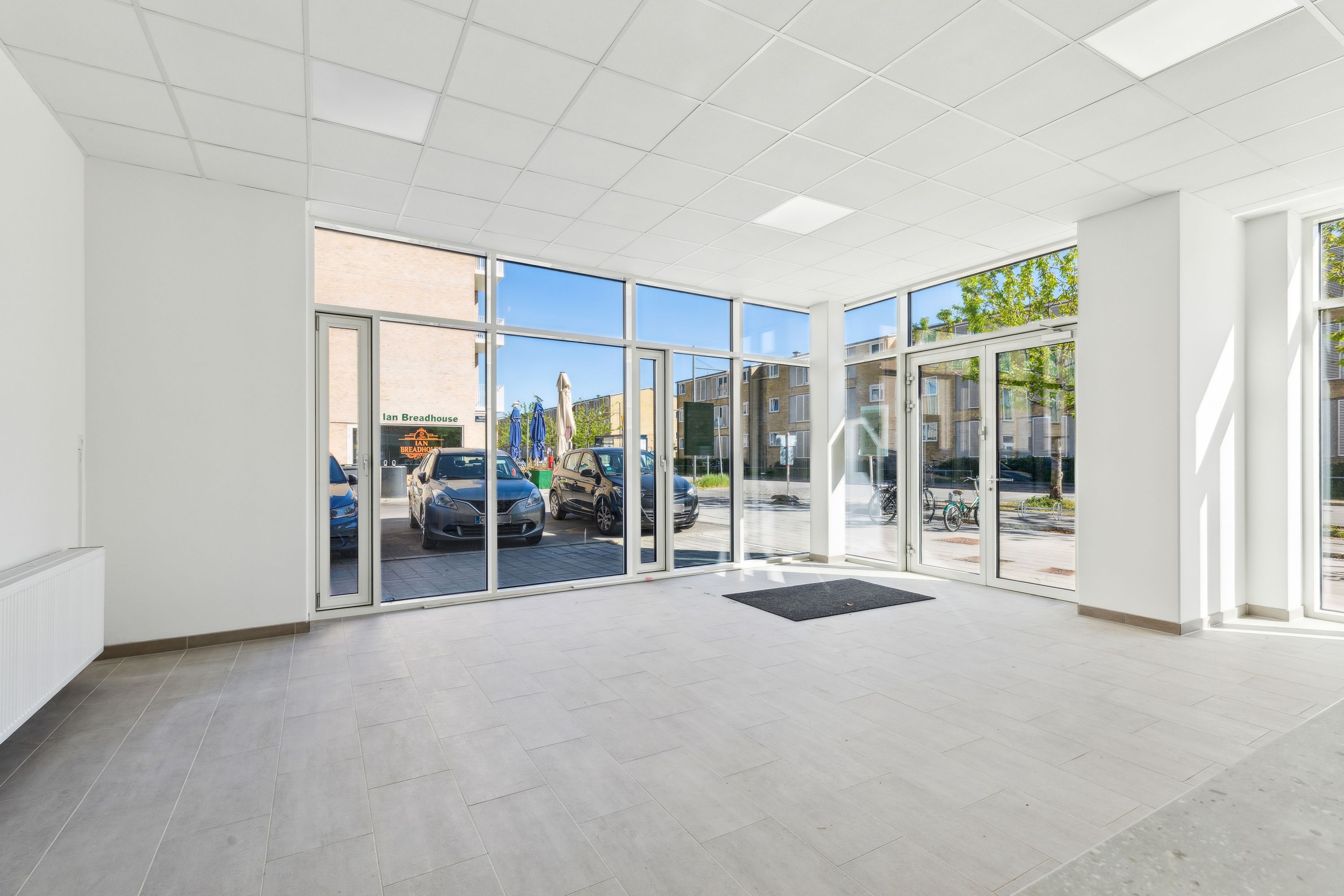 Glasdøre og vinduer med udsigt til gaden, hvor biler parkeret udenfor og grønne træer og buske. Inde i lokalene hvide vægge,