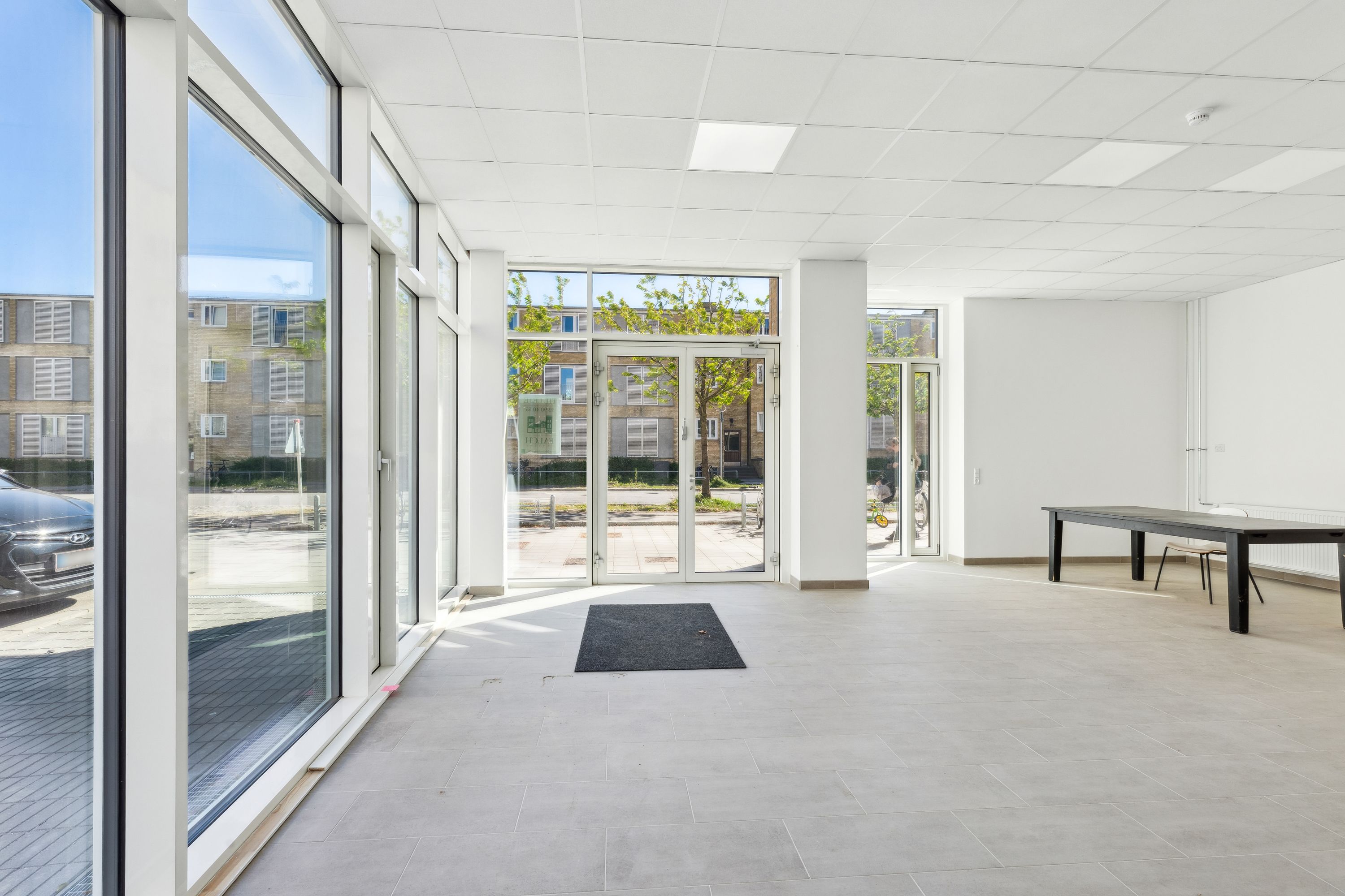 Store glasruder, hvide vægge og lofter samt en stor glasdør med indgangsmåtte og en lang, simpel møbleret borde med stole.