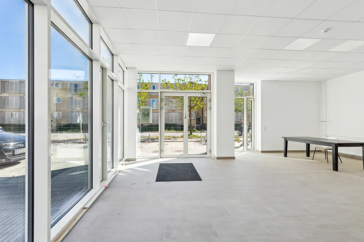 Store glasruder, hvide vægge og lofter samt en stor glasdør med indgangsmåtte og en lang, simpel møbleret borde med stole.