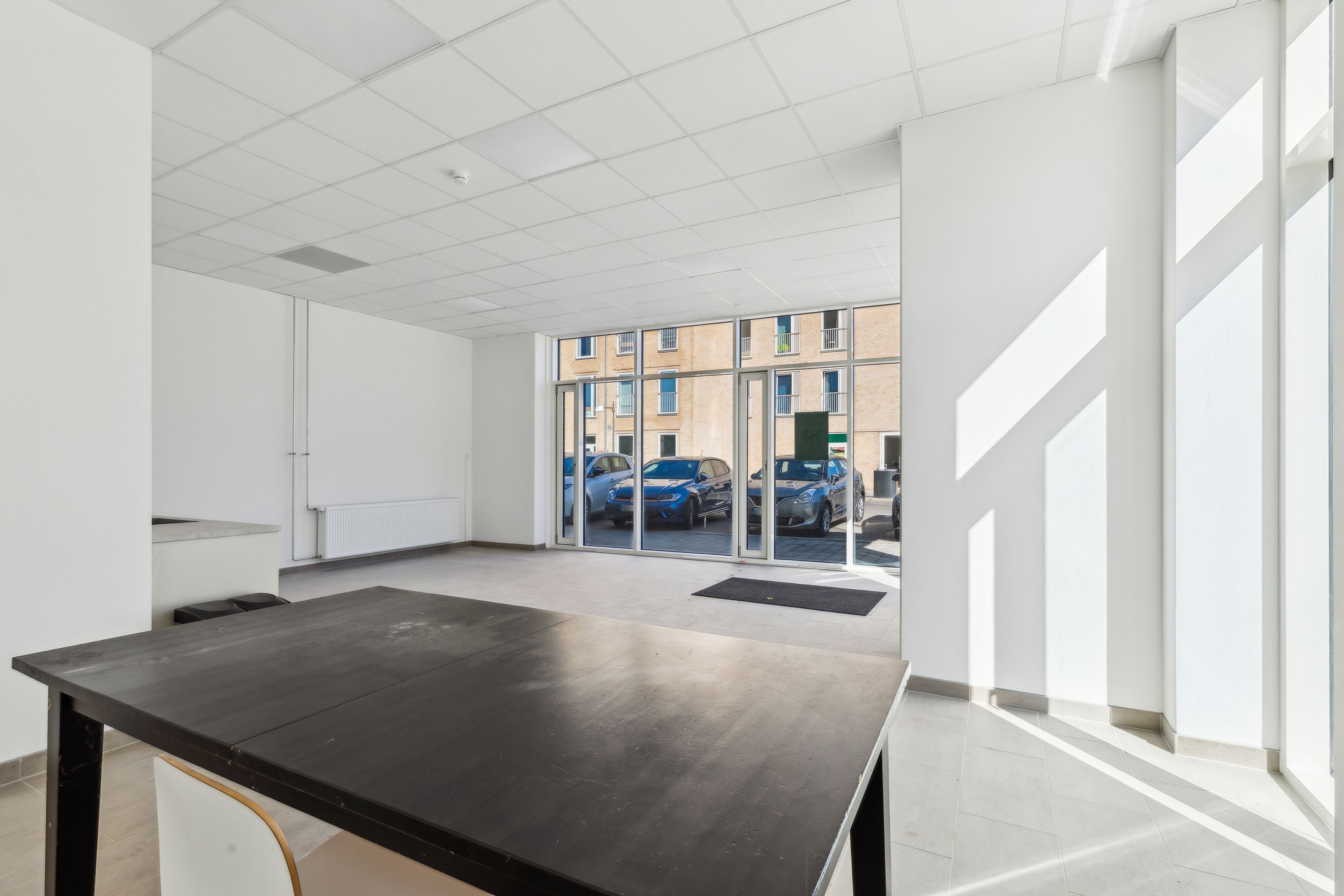 Moderne og åben erhvervslejemål med stort glasvindues og parkeringspladser. Indeholder et træbord og stol samt hvide vægge.