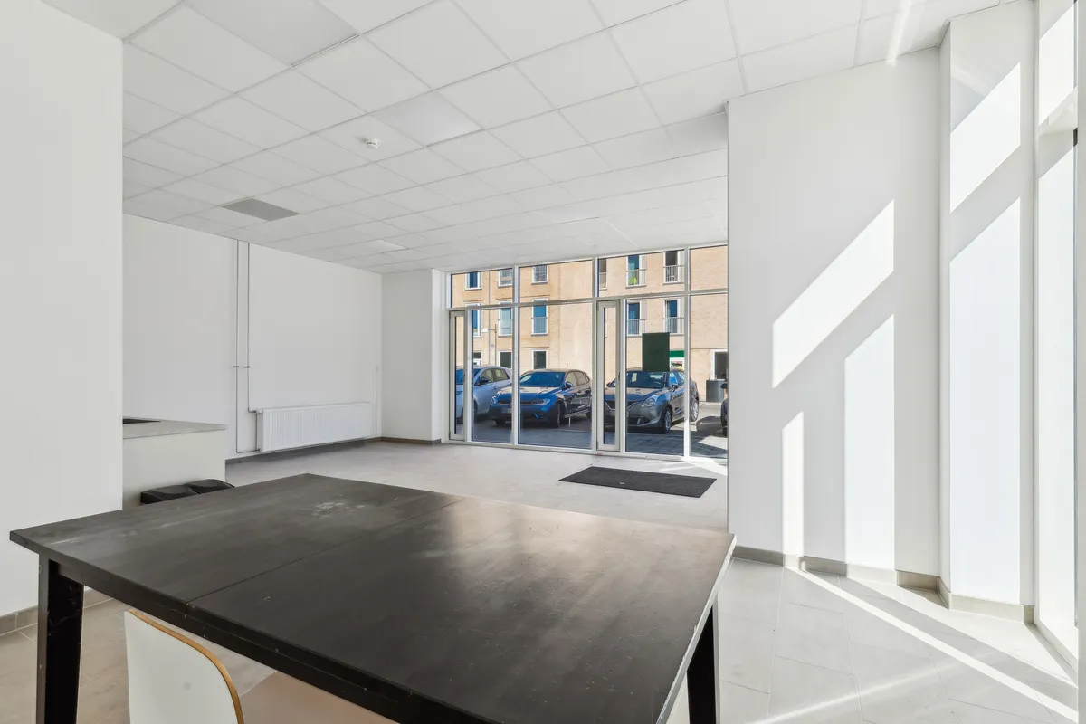 Moderne og åben erhvervslejemål med stort glasvindues og parkeringspladser. Indeholder et træbord og stol samt hvide vægge.