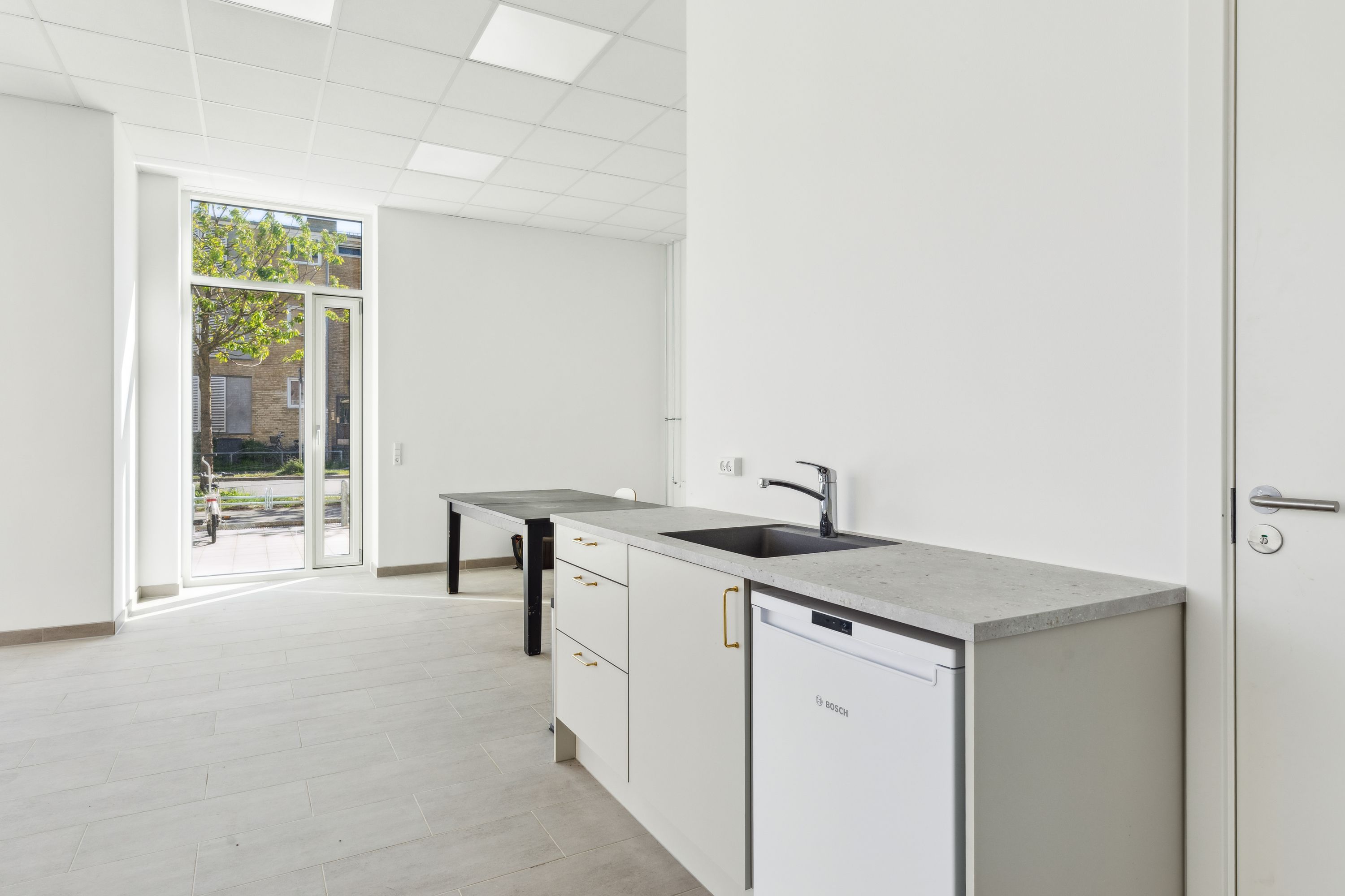 Moderne køkken med hvide vægge, lyse loftspaneler, glasdør, stenkøkkenbord, hvid vaskemaskine og en moderne vandhane