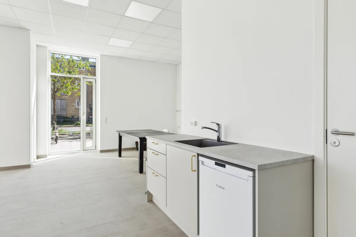 Moderne køkken med hvide vægge, lyse loftspaneler, glasdør, stenkøkkenbord, hvid vaskemaskine og en moderne vandhane