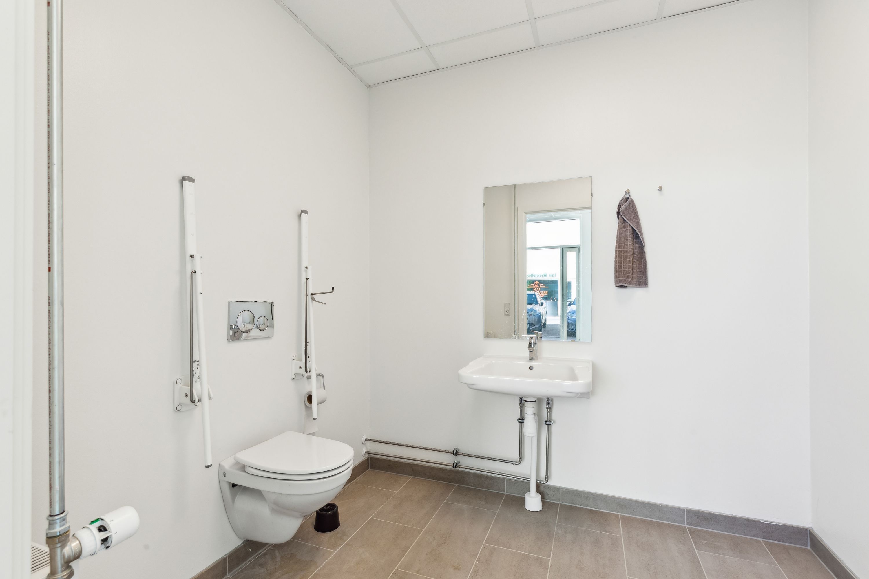 Toilet med håndtak, hvidt toilet med toiletpapir, hvidt vask og spejl med hængende håndklæde, hvidt loft og gulv med mørk