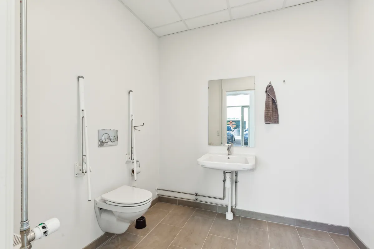 Toilet med håndtak, hvidt toilet med toiletpapir, hvidt vask og spejl med hængende håndklæde, hvidt loft og gulv med mørk