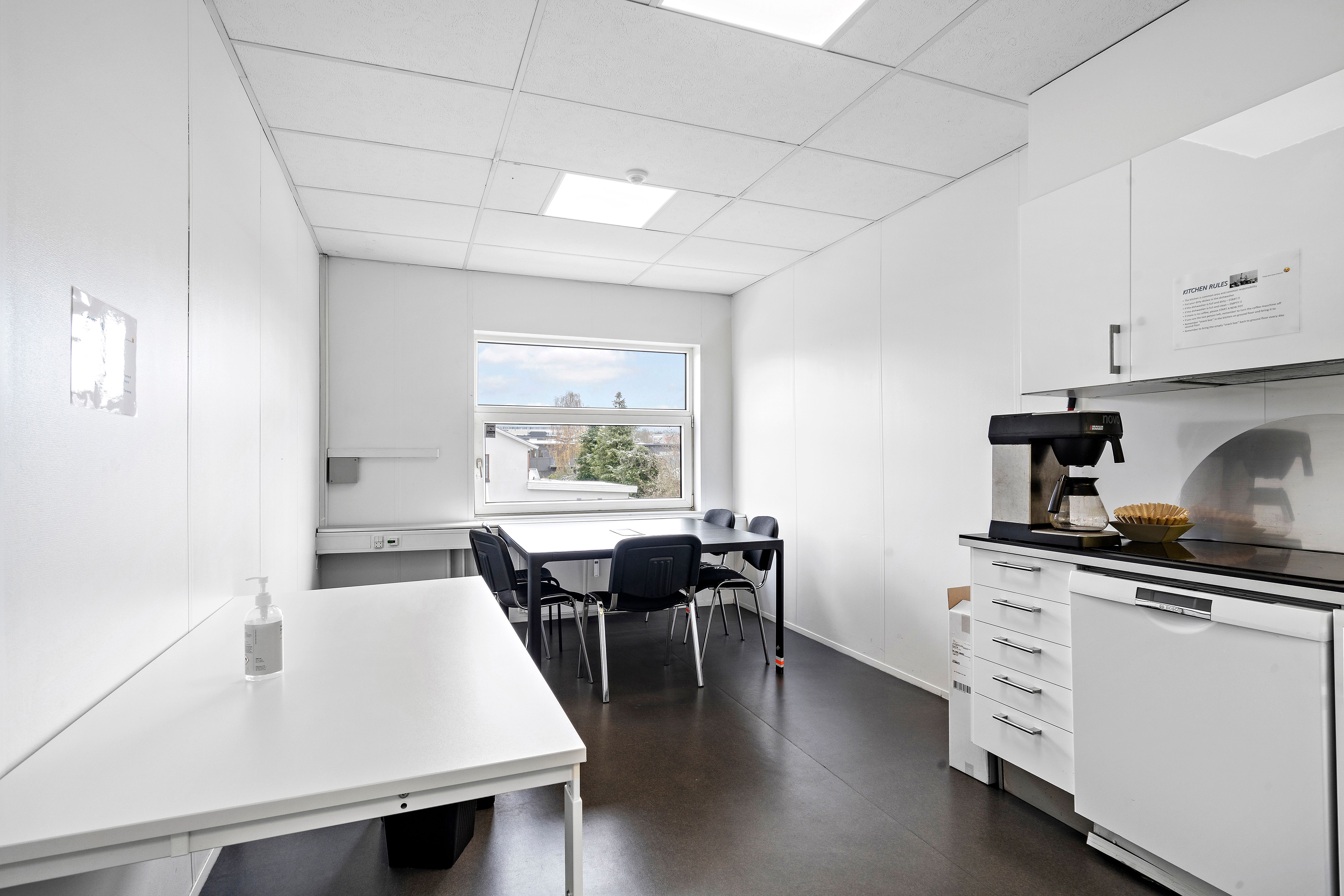 Inde i et lyst og veludstyret kontorrum er der en lang hvid bordplade med en flaske hånddesinfektion og et bord med fem