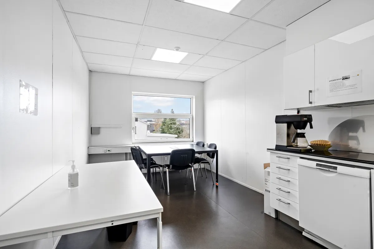 Inde i et lyst og veludstyret kontorrum er der en lang hvid bordplade med en flaske hånddesinfektion og et bord med fem