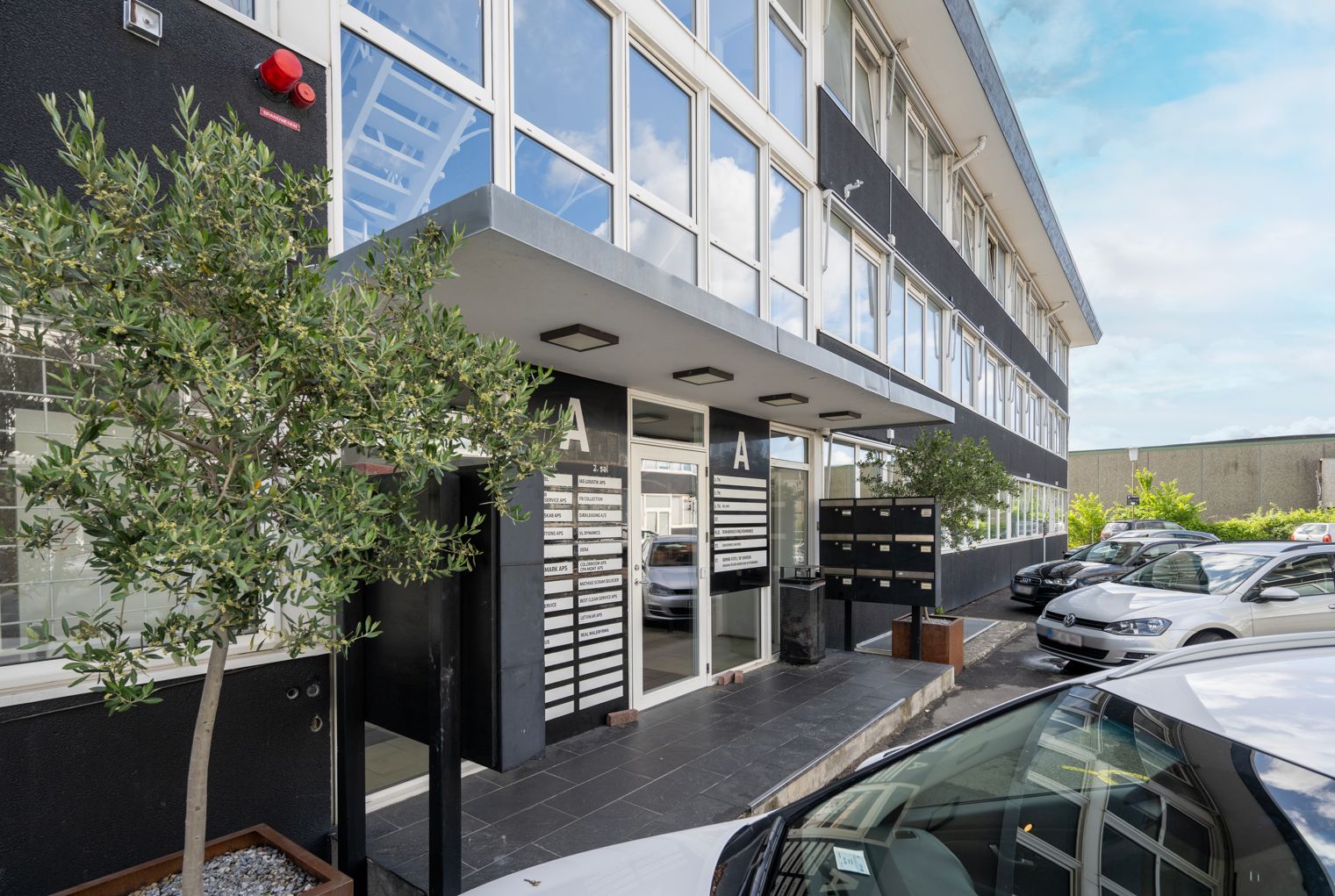 Forretningslokaler med moderne indretning, glasruder, træ og planter samt parkerede biler og postkasser