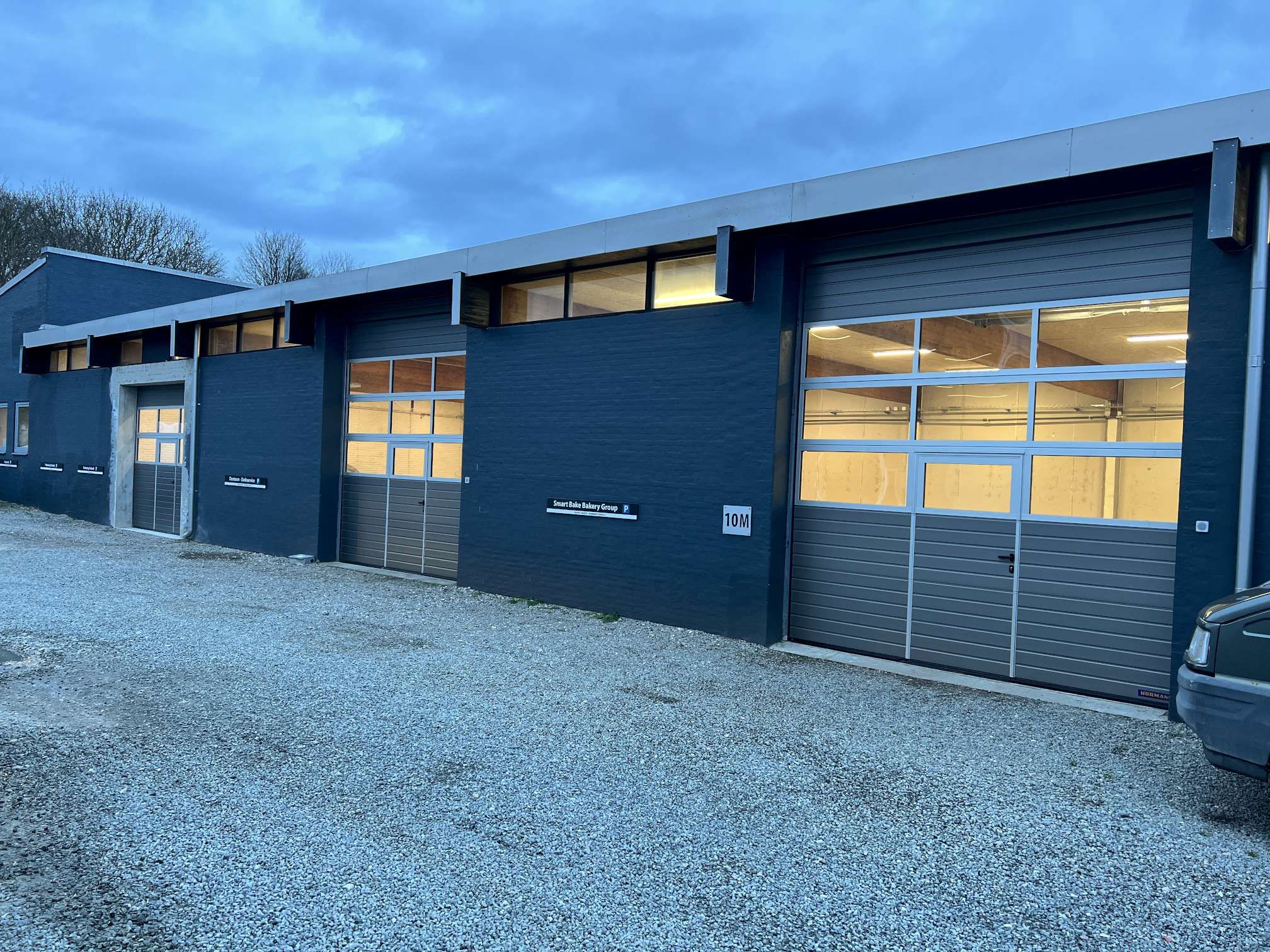 Et blåt lagerhus med tre rulleporte og nummer 10M markeret i højre side. Der er glasruder og døre samt tekst på væggen. En