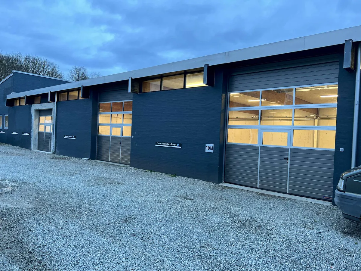 Et blåt lagerhus med tre rulleporte og nummer 10M markeret i højre side. Der er glasruder og døre samt tekst på væggen. En