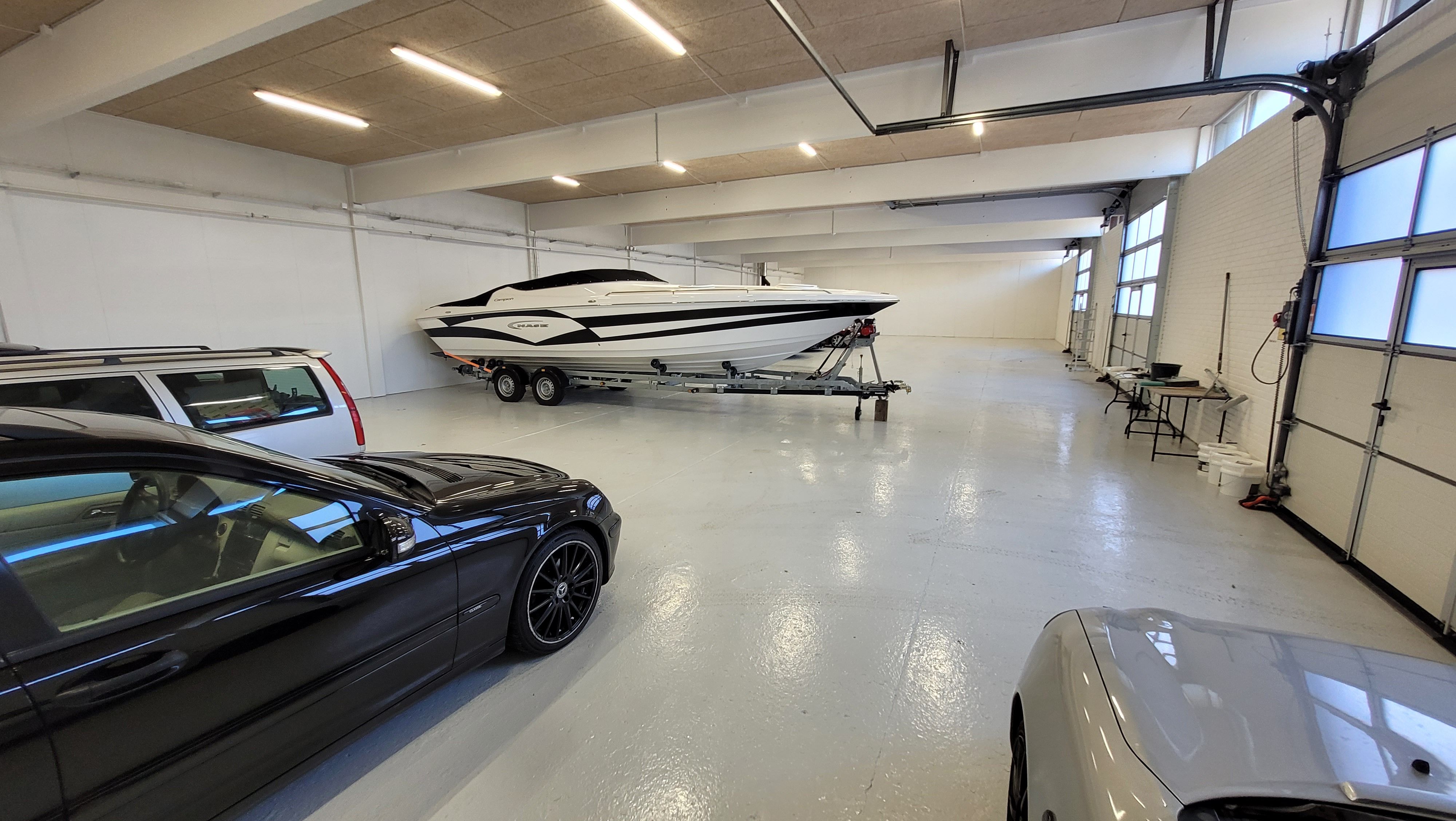Et stort lagerlokale med højt til loftet og hvide vægge indeholder en slæbebåd og biler i garagen