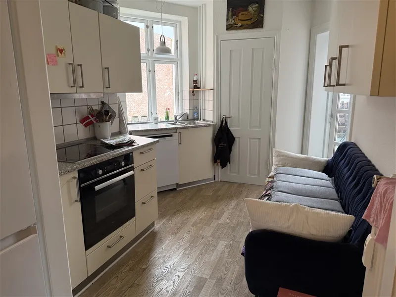 Køkken med trægulv og hvide møbler med en sovesofa og hvidt køleskab i nærheden