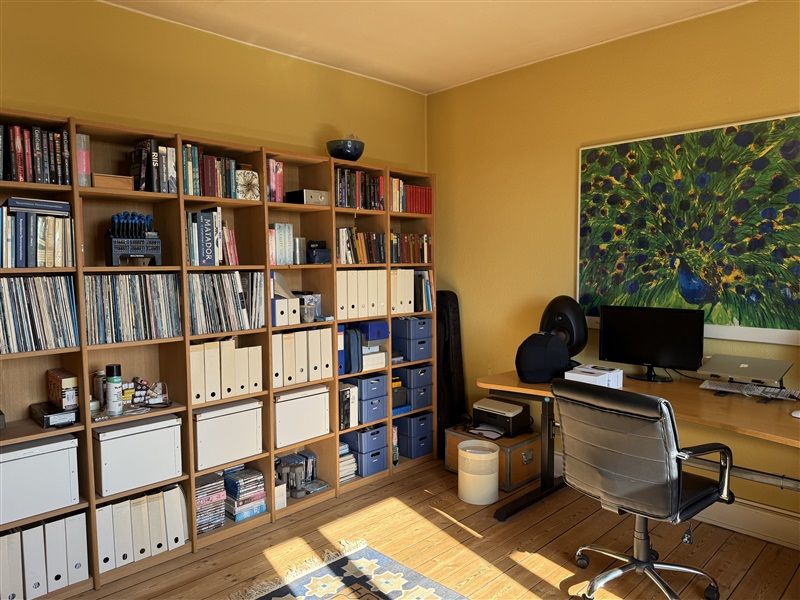 Bibliotek med flere hylder og bøger, dvd'er, mapper og papirer samt en skrivebord med computer og stol