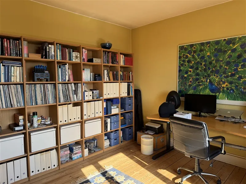 Bibliotek med flere hylder og bøger, dvd'er, mapper og papirer samt en skrivebord med computer og stol