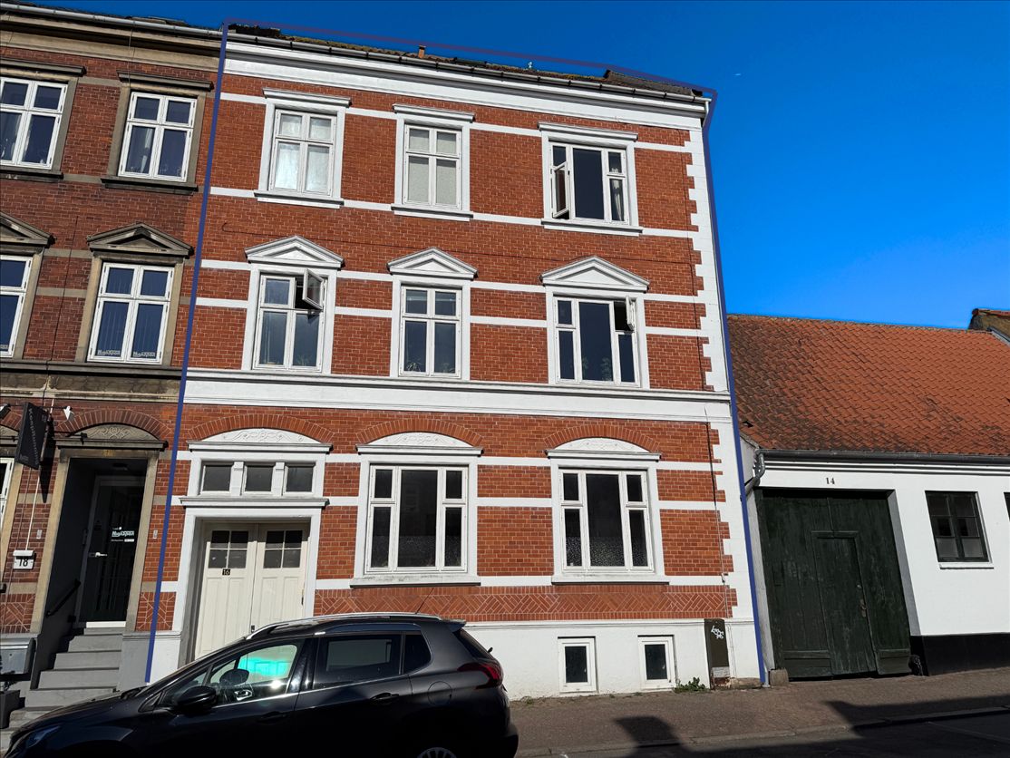 Bygning med røde mursten og hvide vinduesinker, placeret i Fredericia. Sort bil parkeret foran. Skyerne er lyse på himlen.