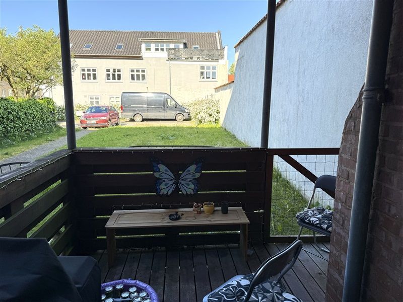 Balkon med udsyn til græsplads med en rødhærdet bil og en vandresti. Bord med potter og papillon.