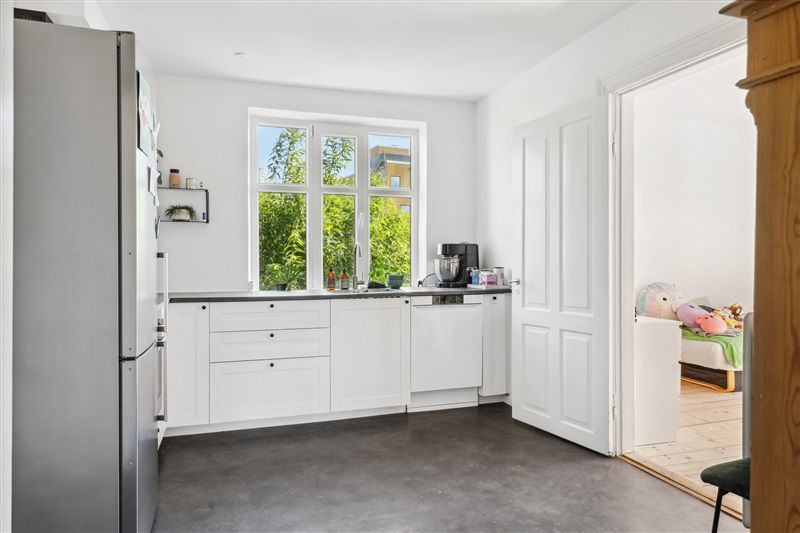 En ren og moderne køkken med hvide vægge, lysegrå gulv, en stor køleskab og en lyse vindue med udsigt til grønne træer