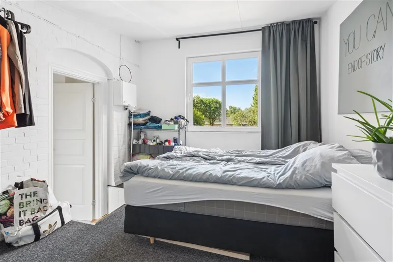 Rent værelse med dobbeltseng og pude i en lejlighed med lyse vægge og gardiner og et skab med tøj