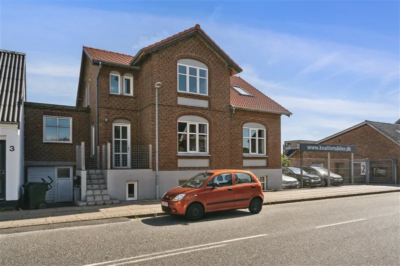 En orange bil parkeret foran et rødt tegltagget hus, som er nummer 3, med flere biler parkeret i baggrunden