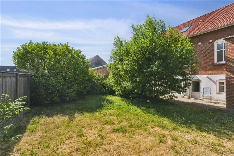 En have med græs, buske og træer omgivet af en høj mur og en bygning med rød tagdækning og lyseruder