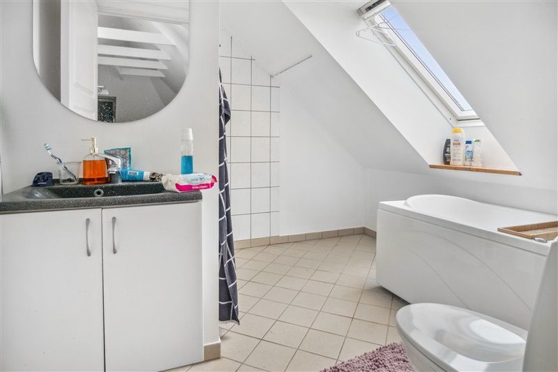 Rent loftsbadeværelse med hvidt badekar, hvidt toilet, spejl og hvide vægge samt badeværelseskabinetter og vindue.