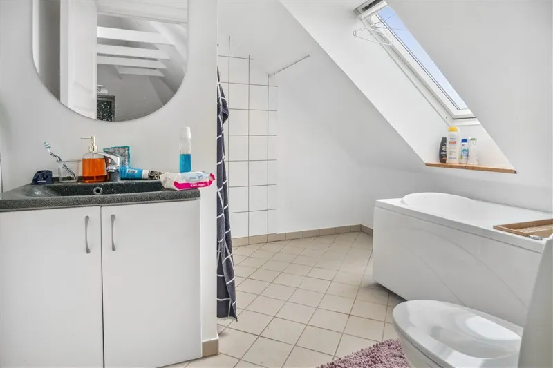 Rent loftsbadeværelse med hvidt badekar, hvidt toilet, spejl og hvide vægge samt badeværelseskabinetter og vindue.