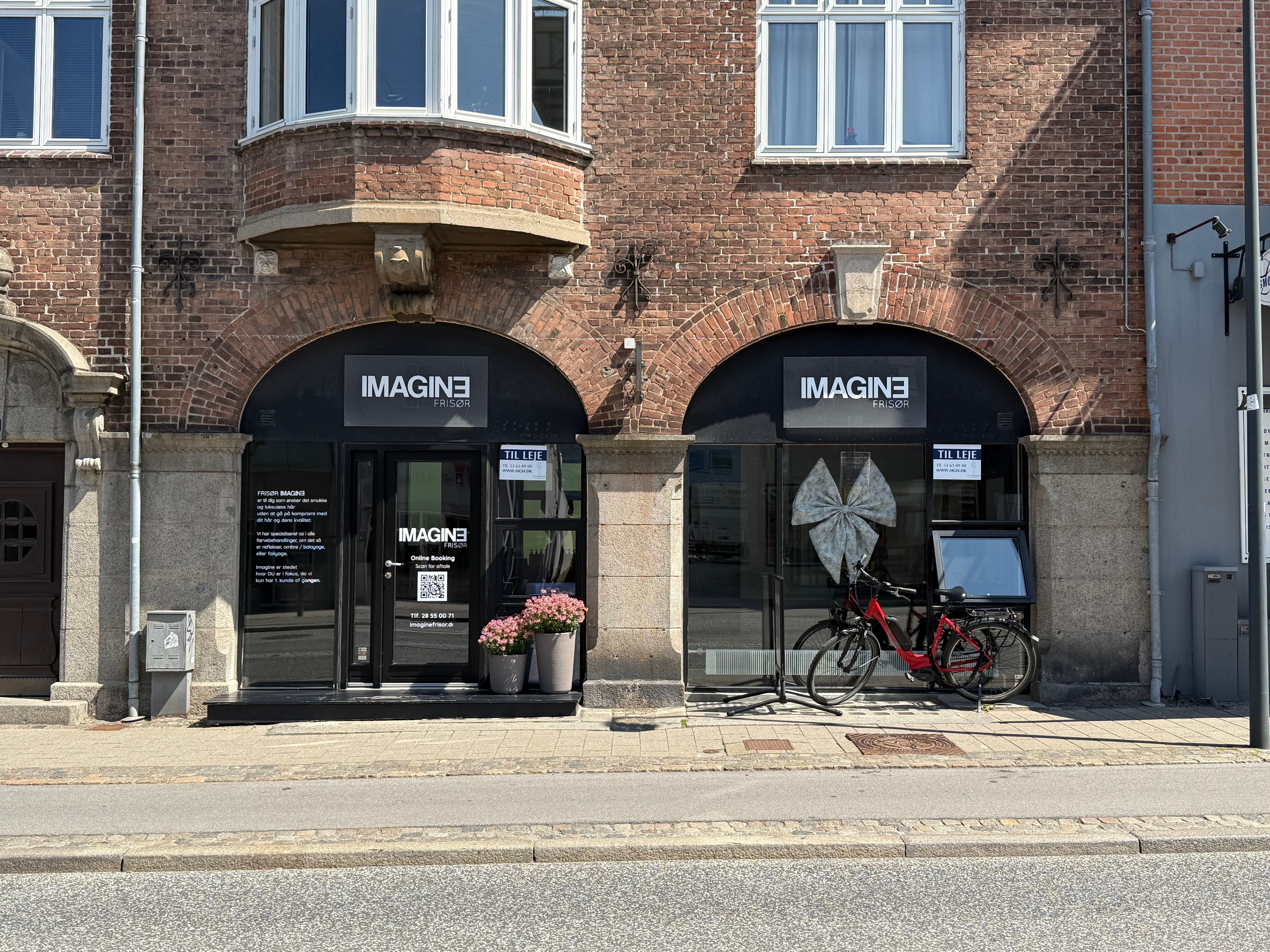To røde cykler står parkeret foran døren til en frisørsalon med navnet Imagine Frisør, hvor der står en QR-kode på døren