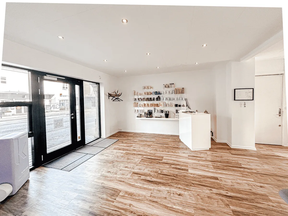 Interiør af et moderne skønhedssalon med højloftede rum og trægulv, hvor en hvid væghylde er fyldt med skønhedsprodukter.